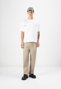 White short-sleeve t-shirt with red text, beige trousers, and black chunky shoes. Model wears a gray baseball cap. Minimalist background.