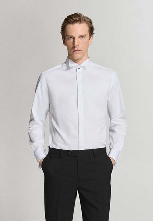 Mann trägt ein weißes Hemd mit verdeckter Knopfleiste und schwarze Anzughose, steht mit den Händen in den Taschen vor schlichtem Hintergrund.