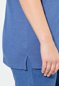 Modrá tričko s uvolněným střihem, které má zakřivený spodní lem a boční rozparky. Materiál vypadá měkký a texturovaný, vhodný pro každodenní nošení.