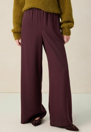 Wijdvallende broek in een diep bordeauxrode stof met een elastische tailleband. Gepaard met donkere schoenen met spitse neuzen en gouden accenten.