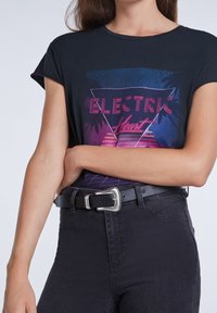 Camiseta negra con un estampado gráfico que presenta "ELECTRIC HEART" en tonos rosa y azul, combinada con unos vaqueros negros de cintura alta y un cinturón con hebilla plateada.