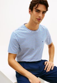 Ριγέ μπλουζάκι σε λευκό και μπλε, με κοντά μανίκια, στρογγυλή λαιμόκοψη και μικρό λογότυπο στο στήθος. Συνδυάζεται με σκούρο μπλε παντελόνι.