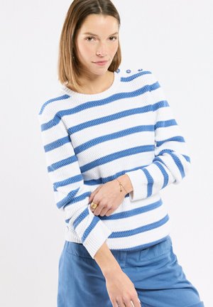 Femme portant un pull blanc à rayures horizontales bleues et un pantalon bleu, ajustant sa manche avec sa main gauche.