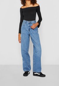 Černý top s dlouhými rukávy odkrytými rameny, spojený s světle modrými vysoko pasovými širokými džíny a černými teniskami s bílými detaily.