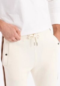 Pantalones de chándal blancos con bolsillos laterales, franjas laterales marrones y cintura con cordón. La tela parece suave y texturizada, adecuada para un uso casual.