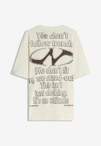 Kermankeltainen, ylivalkoinen t-paita, jossa on rohkea musta teksti selässä graafisilla yksityiskohdilla ja lyhyet hihat. Rentohenkinen puuvillainen malli.