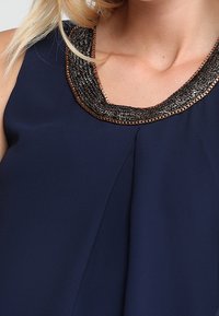 Haut sans manches marine avec un design drapé, mettant en valeur un col orné de métal noir et doré et des plis à l'avant pour une texture supplémentaire.