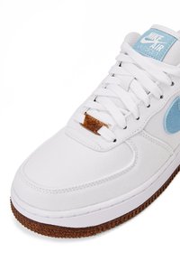 Witte Nike Air Force 1 sneaker met lichte blauwe accenten, bruine getextureerde zool, witte veters en een bruine veterlabel op een witte achtergrond.