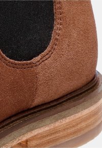 Bottine en suède marron avec panneau latéral élastique noir et semelle en bois stratifiée, montrant des coutures détaillées et une texture.