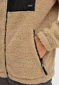 Giacca in pile marrone con accenti in nylon nero, tasca con zip e tasca laterale. Superficie testurizzata con un aspetto morbido e soffice.