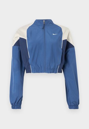 Niebieska kurtka z białymi i granatowymi blokami kolorów, obcięta, z wysokim kołnierzem, przednim zamkiem błyskawicznym, elastycznymi mankietami i błyszczącą fakturą. Logo Nike na piersi.