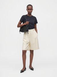 Femme debout portant un t-shirt noir, un short plissé crème longueur genou, des ballerines noires pointues, et tenant un sac à bandoulière noir.