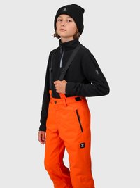 Ung person iført sort hue og jakke, klare orange ski bukser med seler, der står foran en lys grå baggrund.