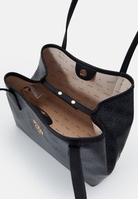 Zwarte en grijze GUESS schoudertas met een open bovenkant die de beige binnenvoering toont, bedrukte GUESS-logo's en twee magnetische drukknoopsluitingen.