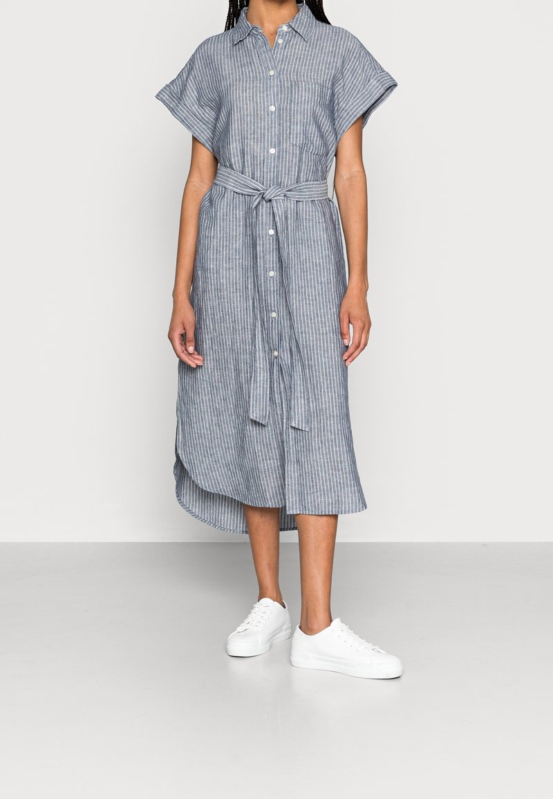 Robe chemise rayée bleue et blanche avec des manches courtes, une ceinture à la taille et une longueur midi, associée à des sneakers blanches.
