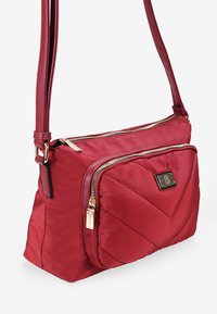 Bolso cruzado de nylon rojo con un diseño de chevrón acolchado, dos bolsillos con cremallera, un acento de hardware en tono dorado y una correa ajustable.