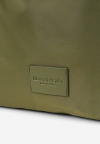 Oliwkowa torba z nylonu z fakturowaną powierzchnią i prostokątną skórzaną metką z wytłoczonym tekstem "Marc O'Polo" oraz "Est. w Sztokholmie."