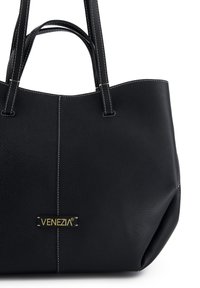 Black leather handbag with textured finish, featuring contrasting white stitching and a gold logo patch on the front. Structured shape with two handles.