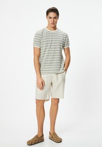 Hvid herre t-shirt med korteærmer i bomuld, dekoreret med grøn stribet mønster, parret med lyse beige linned shorts og brune ruskindssandaler.