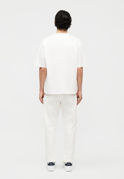Άνδρας με κοντά σγουρά μαλλιά, που φοράει απλό λευκό κοντομάνικο μπλουζάκι, λευκό παντελόνι και λευκά αθλητικά παπούτσια Polo, σε θέα από πίσω.