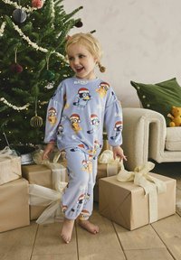Sininen pyjamapuku, jossa on sarjakuvahahmoja jouluhattu päässä, elastiset hihansuut, seistyään paljain jaloin puulattialla käärittyjen lahjojen lähellä.