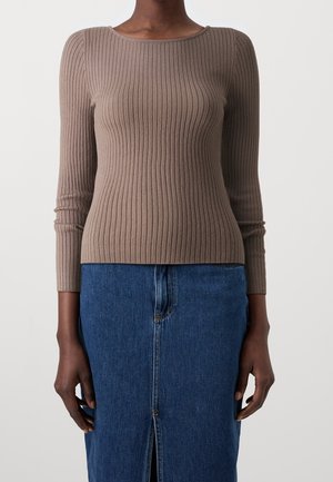 Personne portant un pull ajusté côtelé taupe à manches longues et une jupe en jean bleu avec une fente devant, debout devant un fond uni.