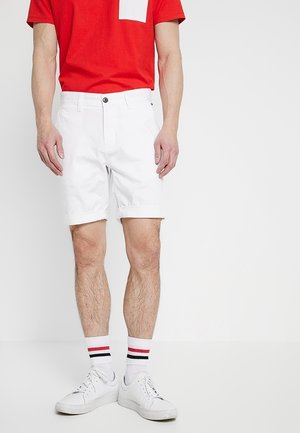 Shorts di cotone bianchi con risvolti, abbinati a una maglietta rossa con una tasca bianca. Scarpe da ginnastica bianche con calzini a righe completano il look.