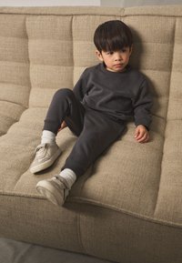 Tamnosivi pulover i trenirka s rebrastim manšetama, nošeni s kremastim tenisicama i bijelim čarapama, sjedeći na teksturiranom bež kauču.