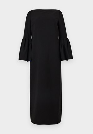 Femme portant une longue robe noire avec de larges manches cloche et des sandales noires à talons hauts, debout devant un fond clair et uni.