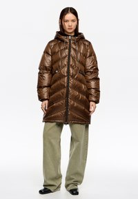 Brązowa kurtka puchowa z kapturem, z pikowaniem w kształcie jodełki, regulowanymi sznurkami i dwiema przednimi kieszeniami na suwak. Noszona na oliwkowo zielonych szerokich spodniach.