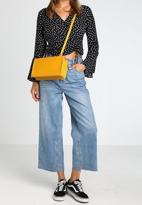 Borsa a tracolla rettangolare gialla con texture liscia, indossata con una maglia nera a pois e jeans a gamba larga azzurri, abbinata a sneaker nere.