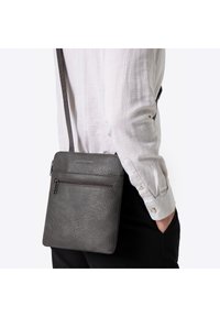 Szara torebka na ramię z teksturowanej skóry, z przednią kieszenią na zamek i regulowanym paskiem, prezentowana na osobie noszącej jasną koszulę.