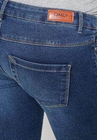 Pantalones vaqueros de mezclilla azul oscuro con una etiqueta de cuero que dice "ONLY". Presenta costuras visibles y un bolsillo trasero con bordes redondeados.