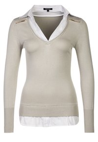 Pull beige clair à manches longues avec un col en V, accompagné d'une chemise blanche à col en dessous et de fermetures éclair dorées sur les épaules.