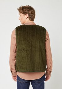 Gilet di velluto a coste verde con righe verticali, caratterizzato da un colletto alto e orlo arrotondato, indossato sopra una camicia a quadri chiara.