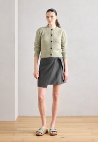 Lichtgroene gebreide cardigan met textuurdesign en zwarte knopen, gecombineerd met een donkergrijze wikkelierskirt en lichtgroene platform sandalen.