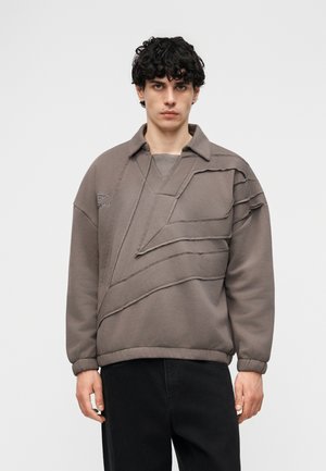 Mladý muž s kučeravými vlasmi má na sebe taupe oversized mikinu s vrstvenými diagonálnymi panelmi z látky a elastickými manžetami, v kombinácii s čiernymi nohavicami.