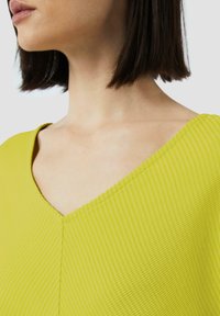Żółta bluzka z prążkowanego materiału o dekolcie w kształcie litery V, wyróżniająca się teksturowanym materiałem i bezszwowym wykończeniem. Skupienie na dekolcie i obszarze ramion.
