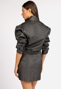 Zwarte textuurjacket met pofmouwen, een taillerende taille en knoopmanchetten, gestyled met een bijpassende strakke rok. Glad, glanzend materiaal.