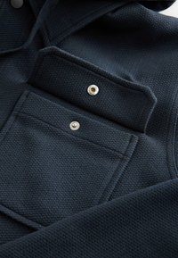 Námořnický texturovaný kabát s náprsní kapsou se stříbrným patentem a hladkým materiálem. Jednoduchý design s jemným vzorem.