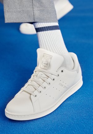 Białe sneakersy Adidas Stan Smith noszone z białymi prążkowanymi skarpetkami z dwoma granatowymi paskami, na tle niebieskiej teksturowanej podłogi.