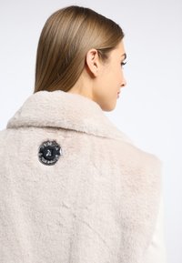 Jasno beżowa kamizelka z futra syntetycznego o miękkiej fakturze, z dużym kołnierzem i dekoracyjnym czarnym guzikiem na plecach z ozdobnym wzorem.