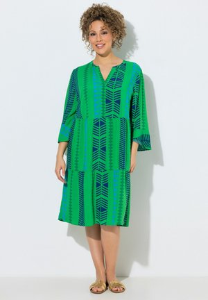 Kobieta o kręconych włosach, ubrana w zieloną sukienkę do kolan z niebieskimi geometrycznymi paskami i beżowymi sandałami, stojąca na białym tle.