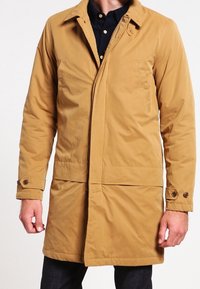 Kakhi isolerad kappa med krage, knappdetaljer på ärmsluten, framfickor och en medellång design. Slät textur med en matt finish.