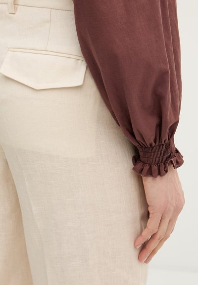 Hand rustend naast beige linnen broek met achterflapzak, gekleed in een lange bruine mouw met gerimpelde manchet en volantdetail.