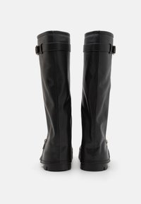 Botas negras hasta la rodilla hechas de cuero sintético suave. Las características incluyen punta redonda, acentos de correa en la parte superior y una suela plana y rugosa.