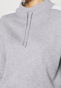 Grijze pullover met een hoge kraag, voorzien van een textuurstof, trekkoorden en witte gestreepte accenten op de schouders.