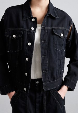 Person iført sort denimjakke med sølvknapper over hvid ribstrikket top, hænder i lommerne på matchende sorte bukser, grå baggrund.