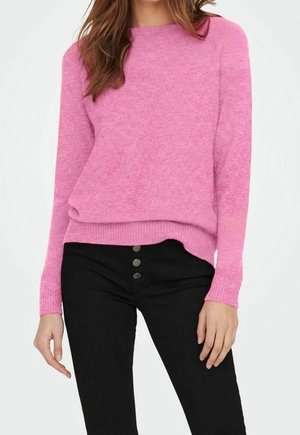Femme portant un pull rose à manches longues et un pantalon noir taille haute avec trois boutons visibles, debout avec une main détendue.