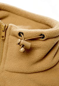 Tissu polaire beige avec une fermeture éclair et un cordon avec un bouton-pression en plastique pour une fermeture réglable.